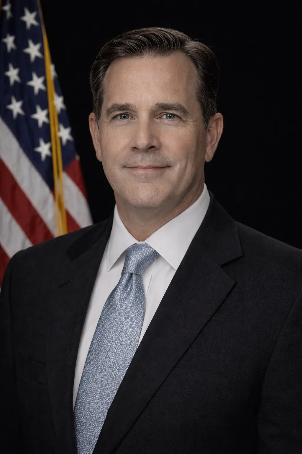 A man in a suit and tie stands before a black background and an American flag.