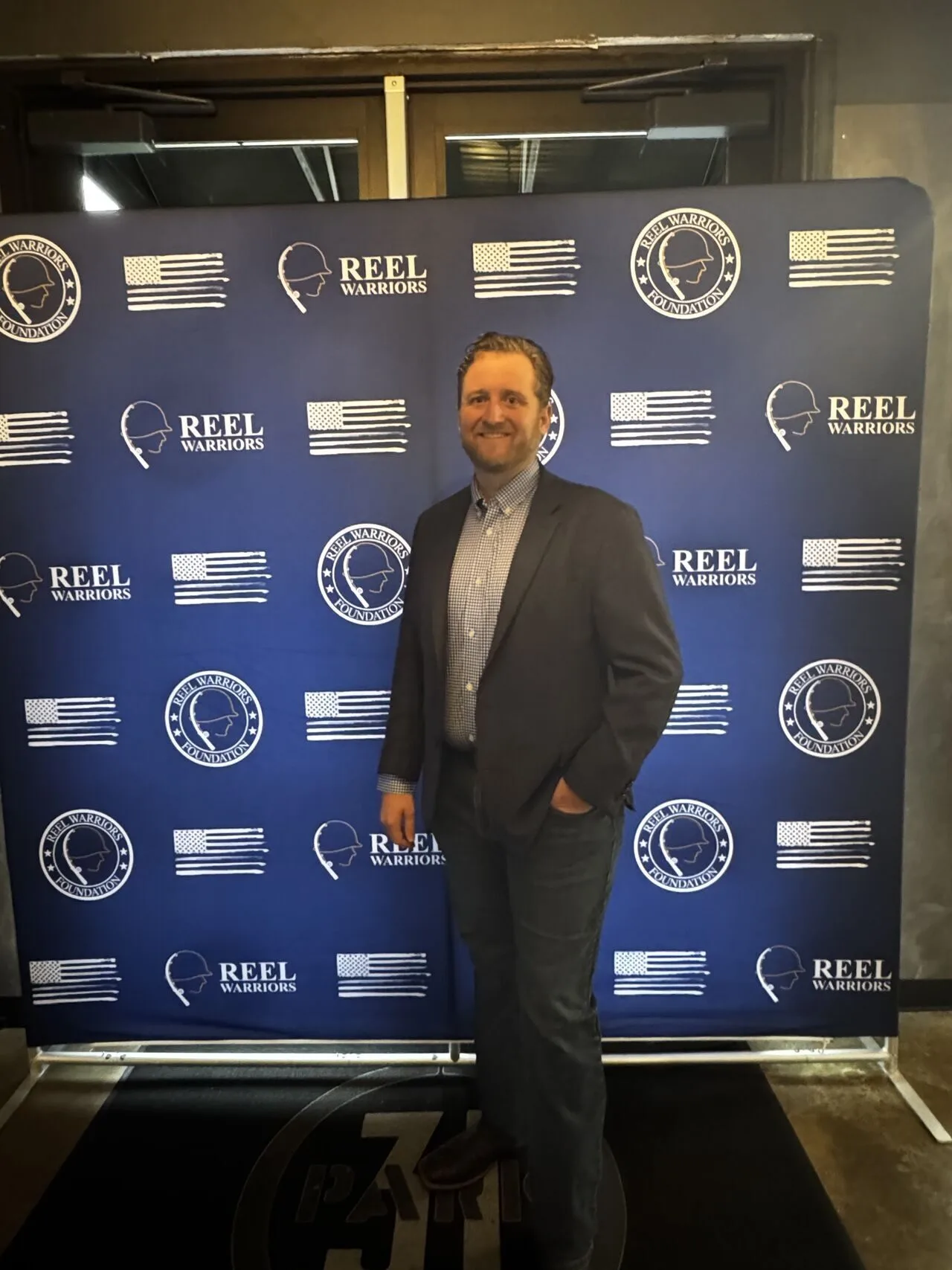 Man in a suit standing in front of a blue backdrop with white logos and flags.