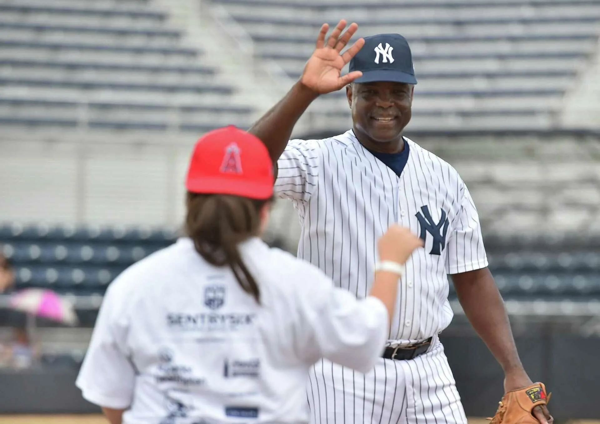 Man in Yankees uniform high-fiving a girl in a red cap and white shirt on a baseball field.
