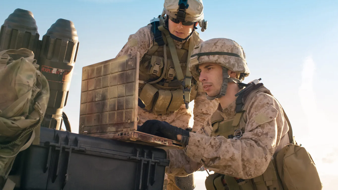 Two soldiers in desert camouflage gear examine a device on a black case outdoors.