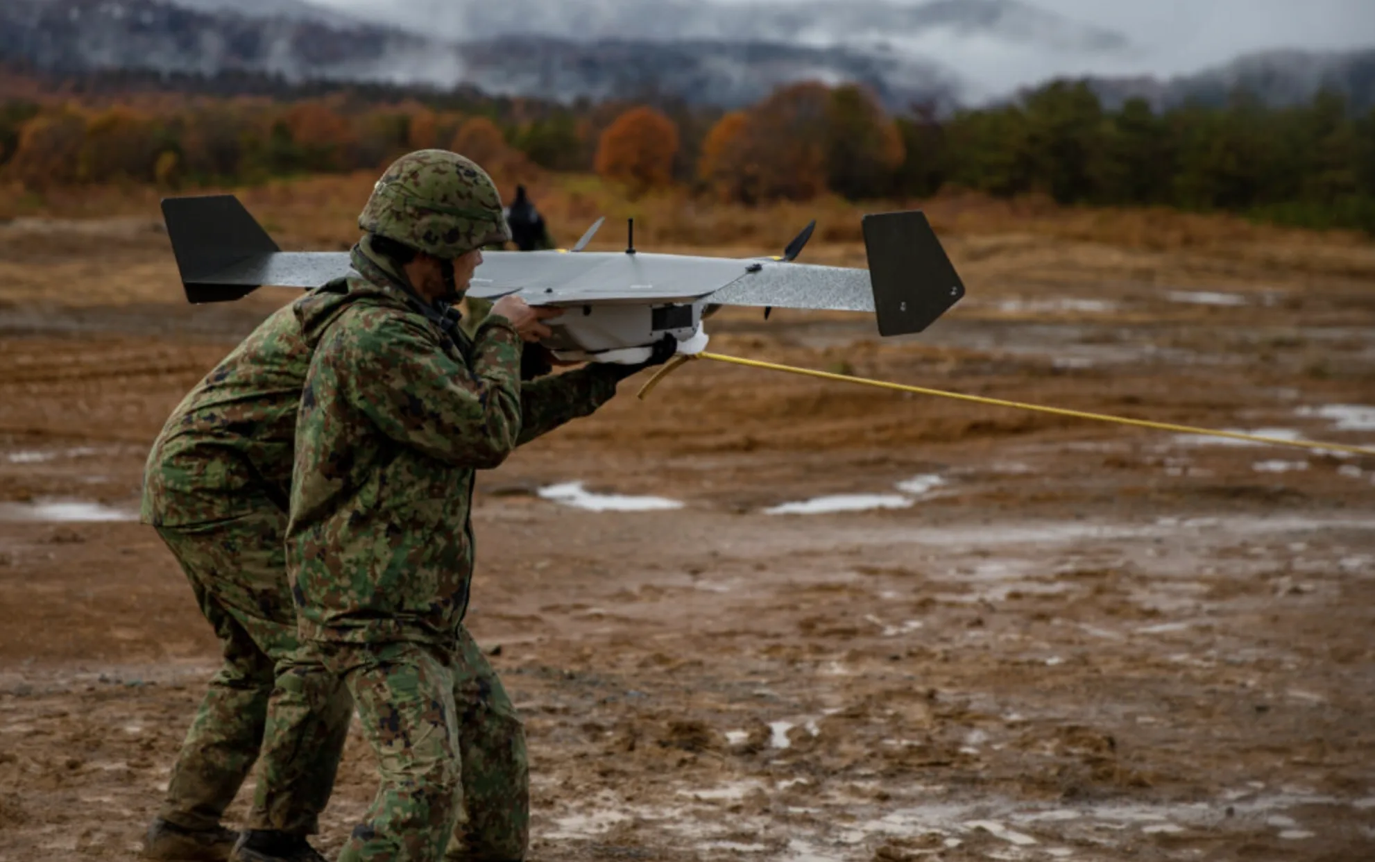 Soldier in camouflage gear launching drone from a dirt field.