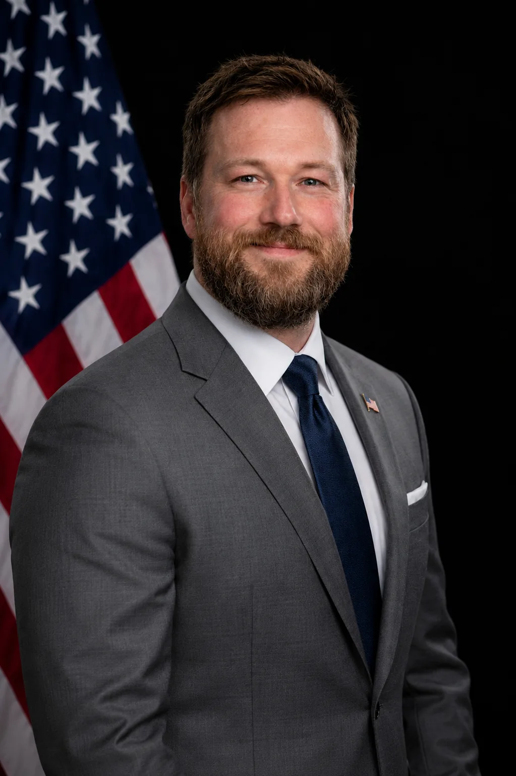 Man in a gray suit with a white shirt and blue tie standing in front of an American flag.