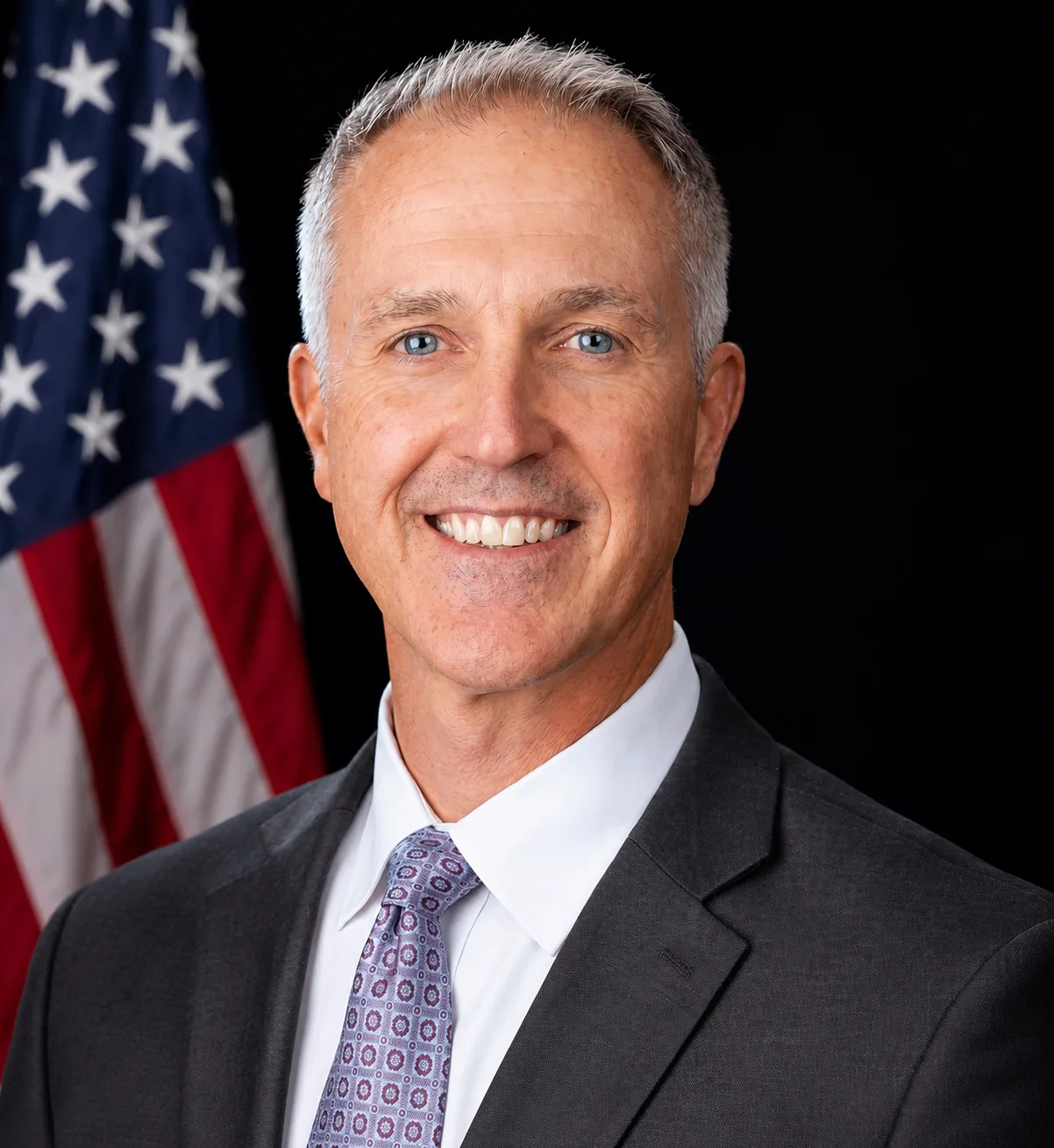 A man in a suit and tie smiles in front of an American flag.
