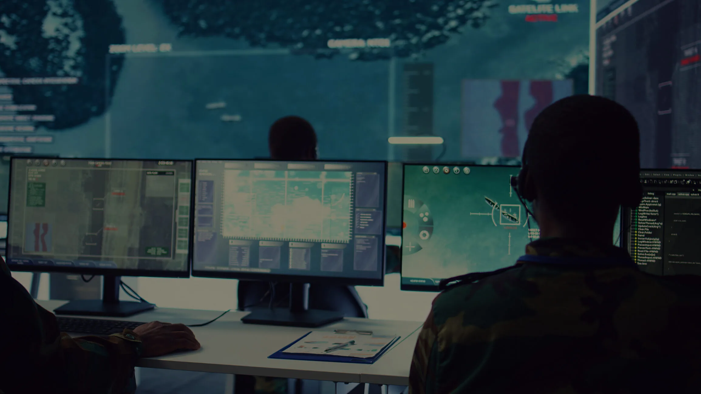Soldiers sitting at a desk with multiple computer monitors displaying various screens and graphics.
