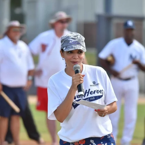 Woman speaks into a microphone on a baseball field with people in the background.